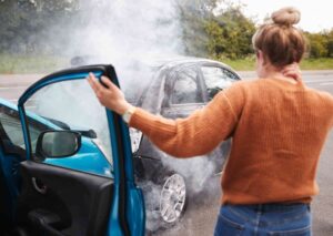 Person holding their neck beside a blue car after a collision with a black car, smoke rising from the engine on a roadside.